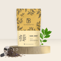 BRU Premium Earl Grey Schwarztee mit natürlichem Bergamotte-Öl im Standbeutel, daneben loser Tee im Teesieb und ein frischer Teezweig